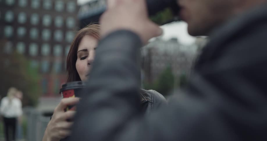 A woman in a leather jacket smiles while drinking coffee and chatting with a man outside in a city