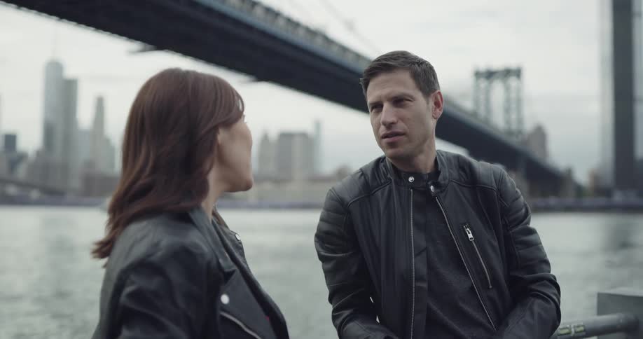 A woman and man in leather jackets chatting while leaning against a handrail beside a river through a metropolis