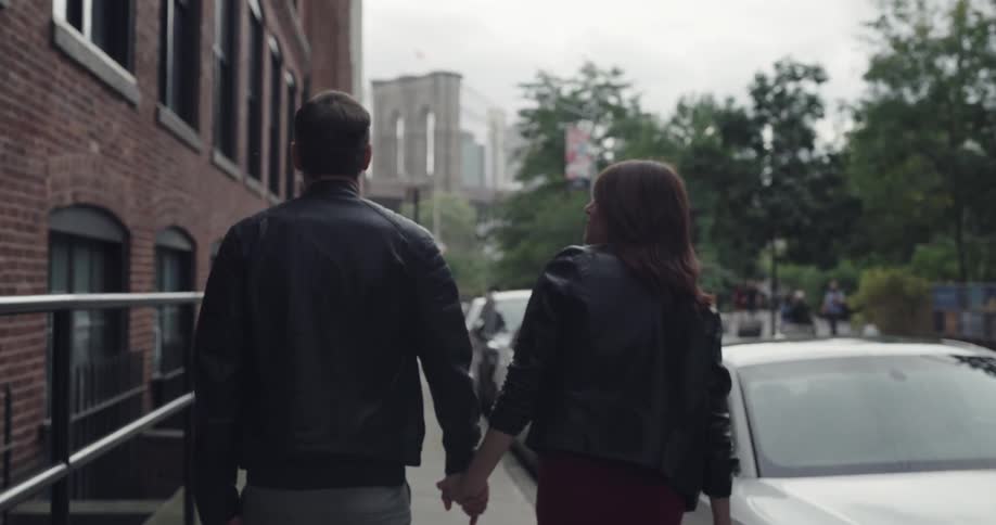 Couple walk along sidewalk between building and parked cars during the day