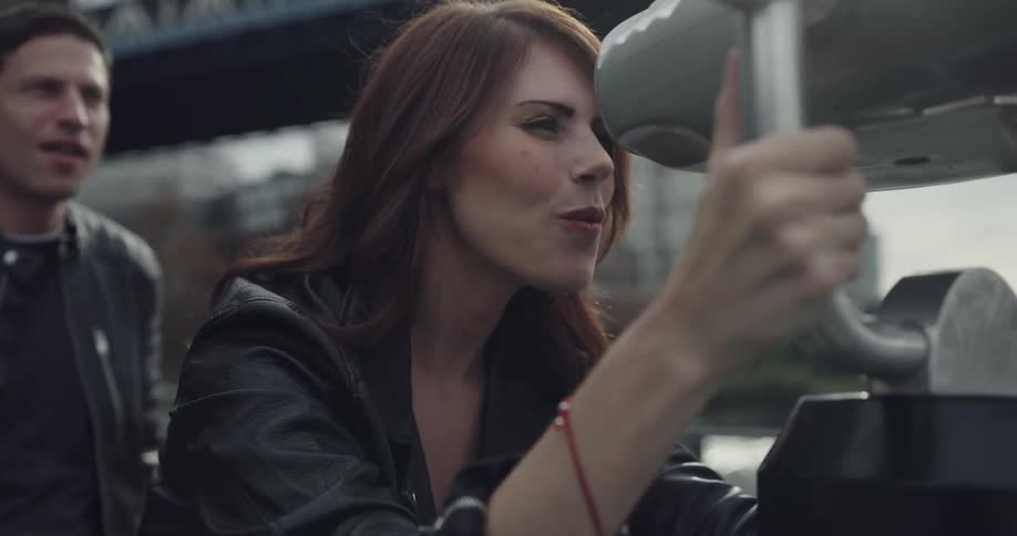 A man playfully blocks the view of a woman who is looking through a set of mounted binoculars at a large city off in the distance