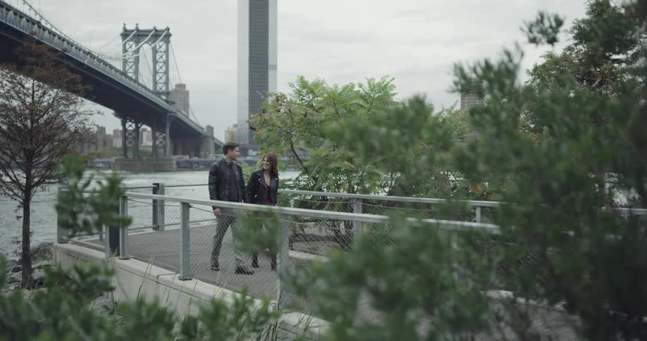 Man and woman holding hands walk along on the promenade next to the river across manhattan bridge and new york city on a cloudy day