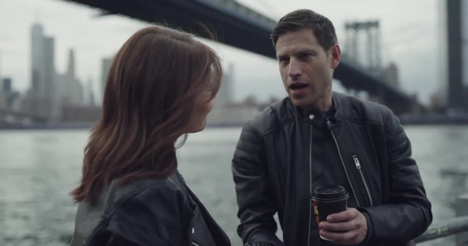 A man and woman who are both wearing leather jackets stand next to a bay across from a large metropolitan city