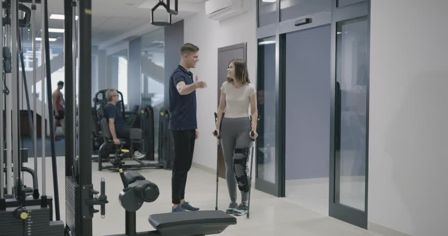 A man greets a woman that is using crutches and a leg brace when she enters a gym and then walks with her