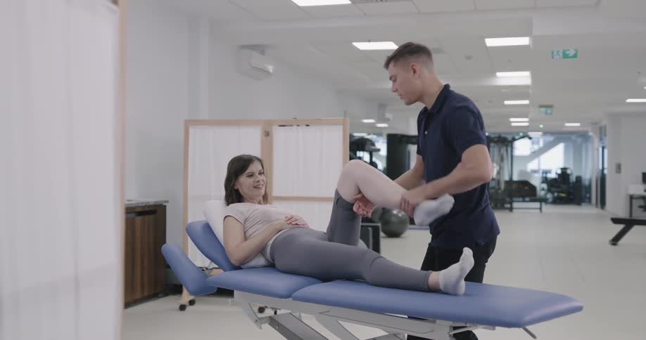 A man stretches a womans leg and pushes on her knee as she lies on a bed