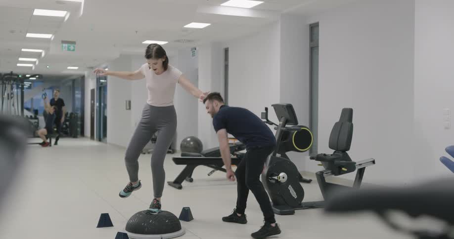A trainer watches as a woman balances on a half ball on one leg and toe taps cones