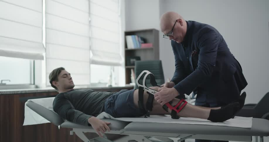A doctor removes a knee brace from a male patient lying on an exam table