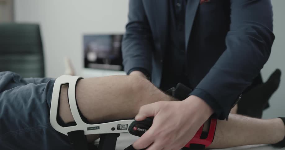 A man is laying on an examination table and has the red and white leg brace removed from around his knee my a man in a suit coat