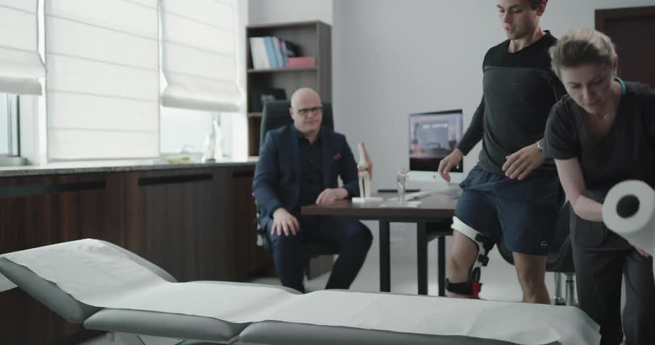 A patient with a knee brace talks to a doctor and lies on a bed for inspection