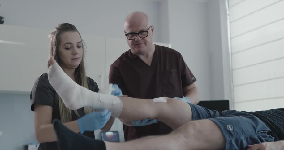 Two medical professionals are working together to remove a bandage from the right leg of a male patient