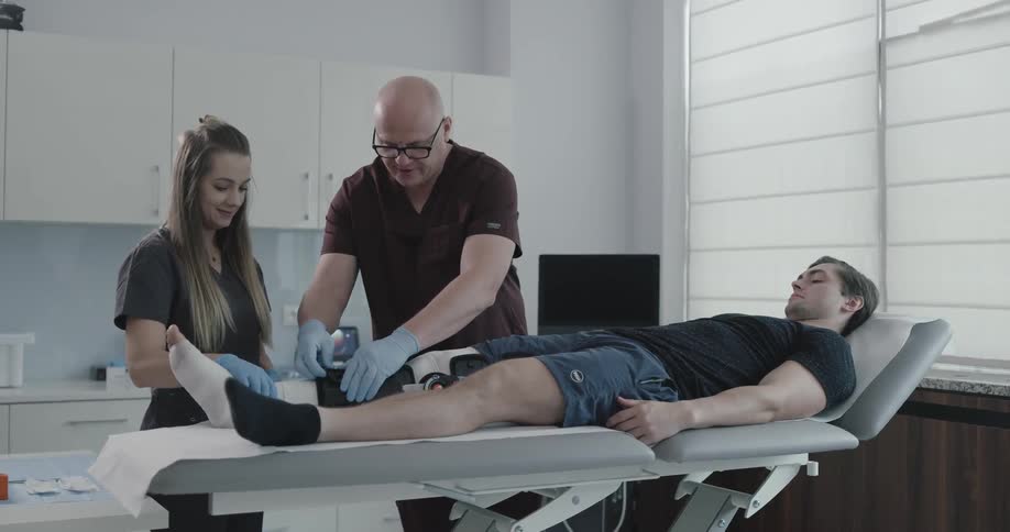 A nurse helps a doctor remove a brace from around a patients wrapped leg in an exam room