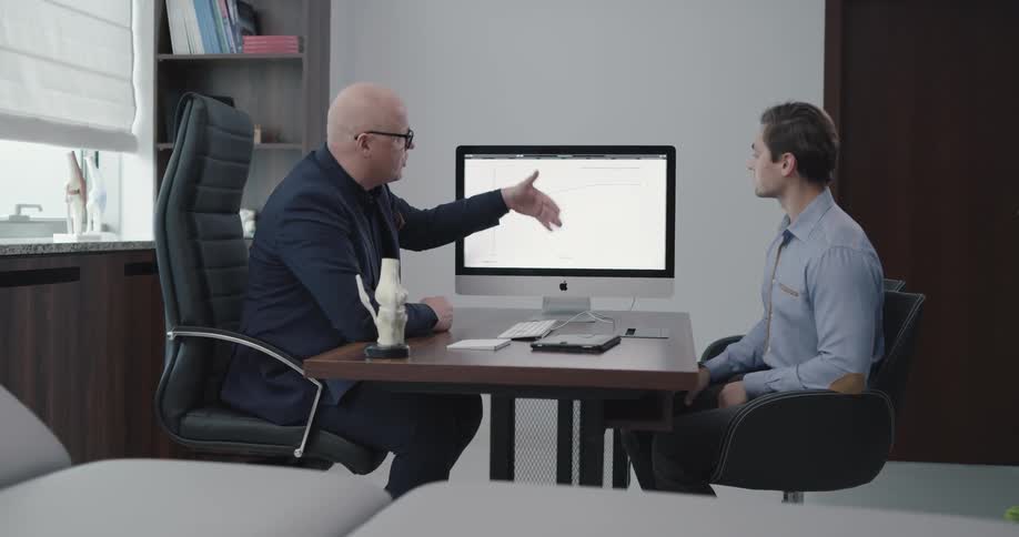 Two men sit at a desk and discuss information on a computer screen