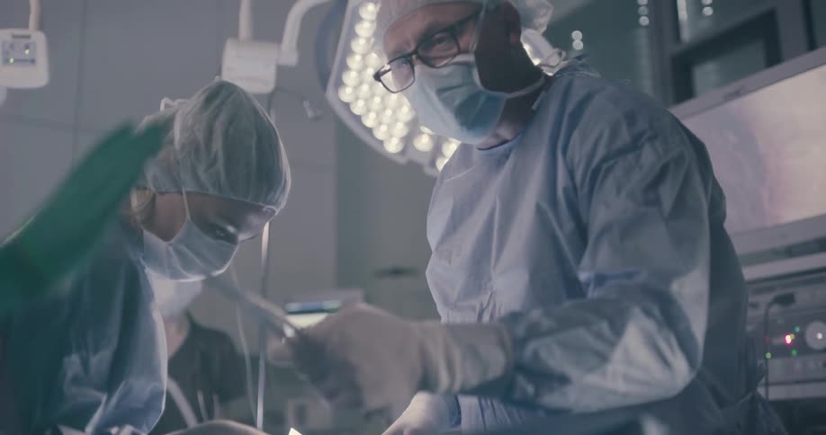 A surgeon reaches his hands out and an assistant gives him a tool to work on a patient