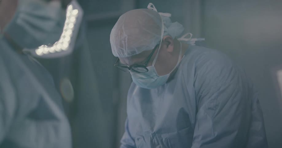 A surgeon wearing a surgical mask and glasses concentrates on a patient as their assistant hands them surgical instruments