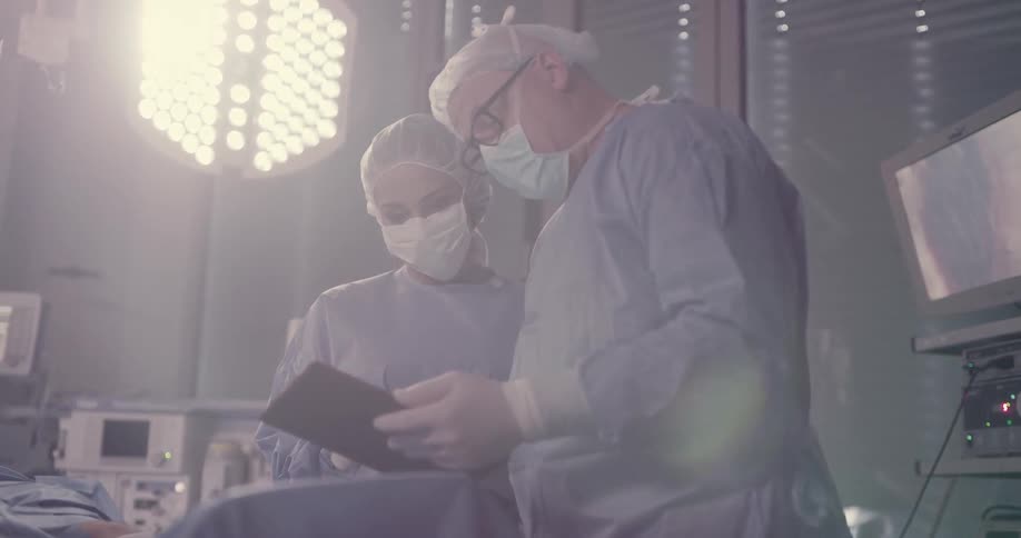 Two surgeons with face masks talk while standing over a patient on an operating table