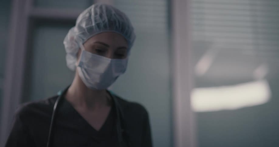 A nurse wearing a mask hair cover and scrubs looks up and leaves the room
