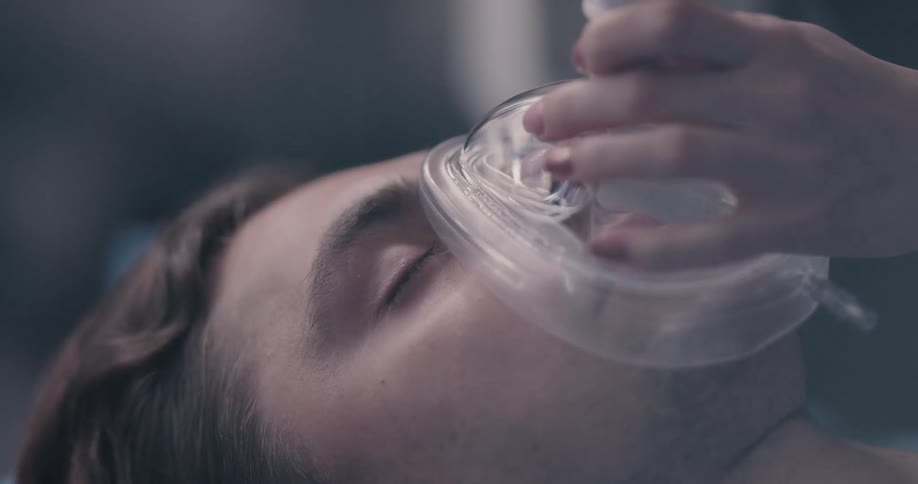 A man is laying down with his eyes closed and a woman puts an oxygen mask on his face
