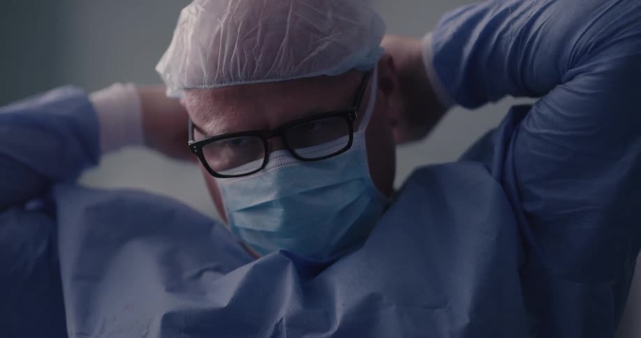 A man wearing a disposable surgical gown and surgical cap ties the strings on a surgical mask around the back of his head