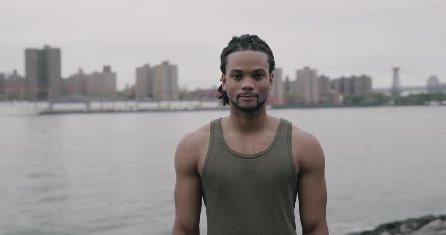 A man standing near the water with the city behind him looks up and smiles