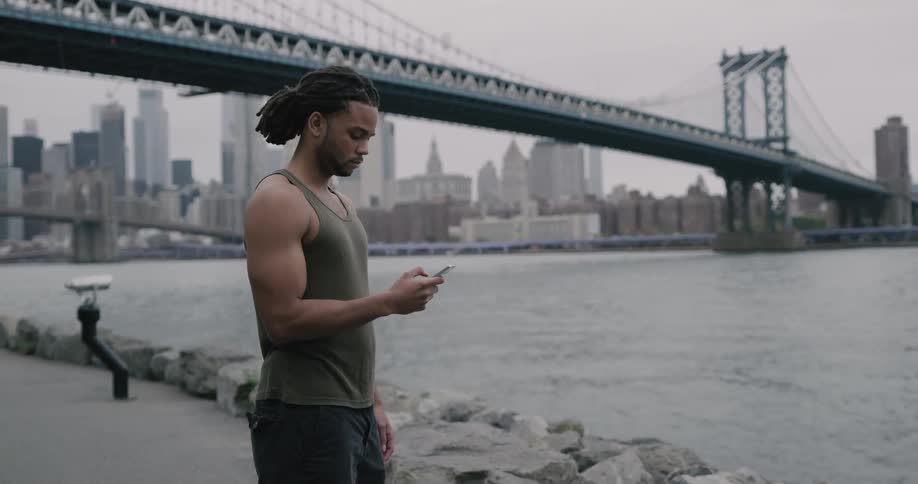 Black man stands and uses smartphone on the promenade next to the river across new york city and brooklyn bridge on a cloudy day