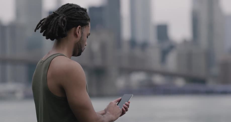 Black man wearing green undershirt uses smart phone by the seaside in the city on a cloudy day