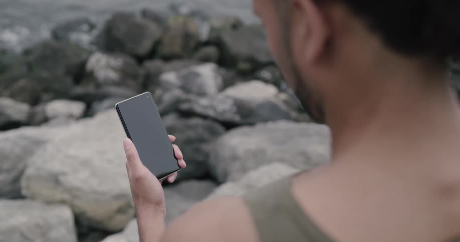 A man standing at the rocky shore of a river looks down at his smart phone in his hand before putting it away