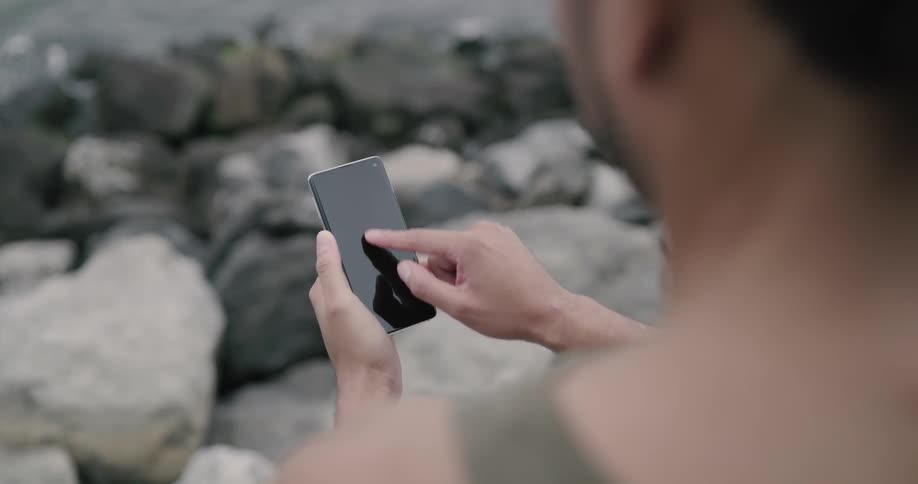 A person sits next to the water on a rocky shore swiping all over their phone