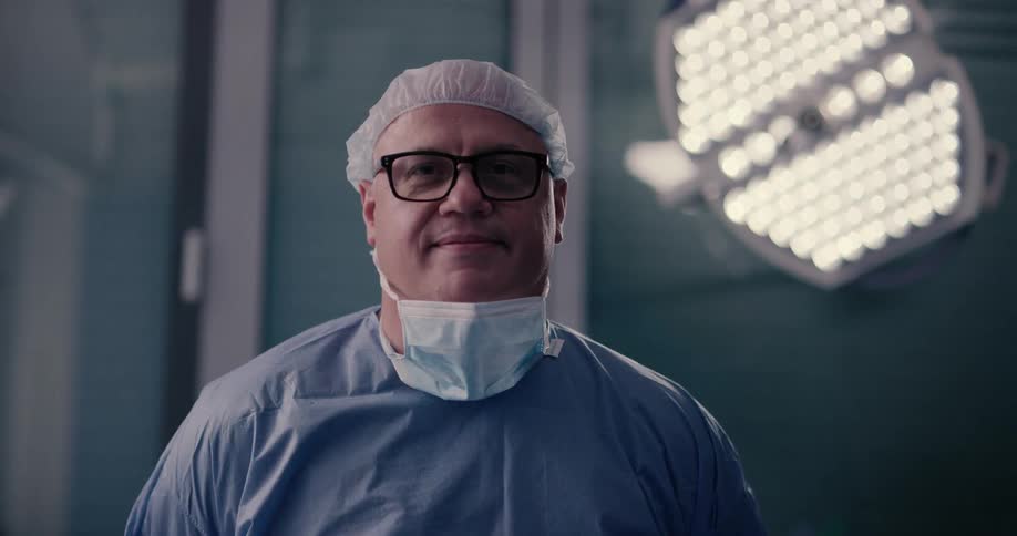 A surgeon removes his face mask while standing in an operating room