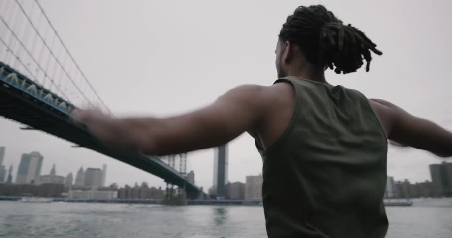 A muscular man jogs along the waterfront adjacent to a bridge and he begins to do jumping jacks as he looks out over the water