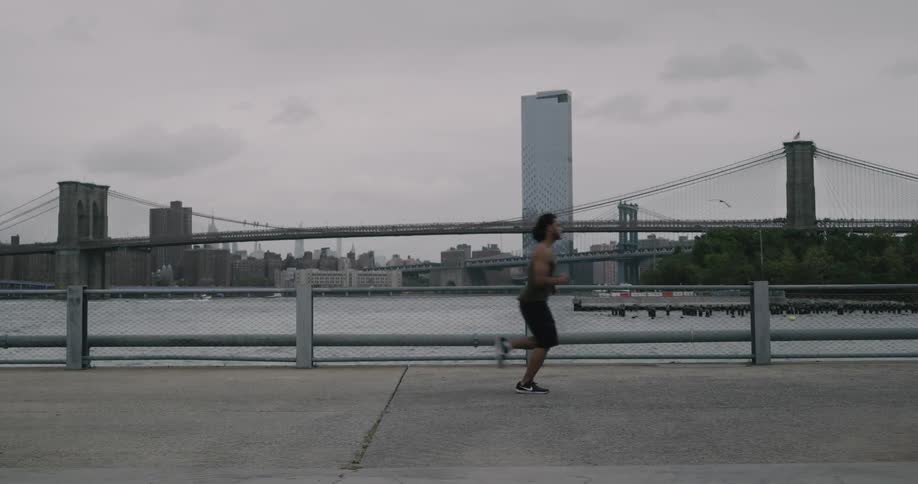 Black man runs along on the promenade and next to the river across the new york city and brooklyn bridge on a cloudy day