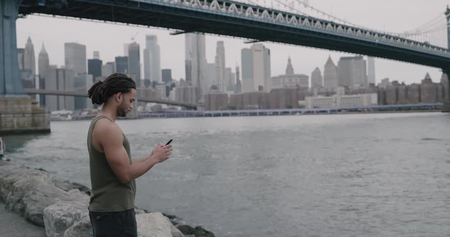 Black man stands on the promenade and uses smartphone next to the river across the new york city and brooklyn bridge on a cloudy day