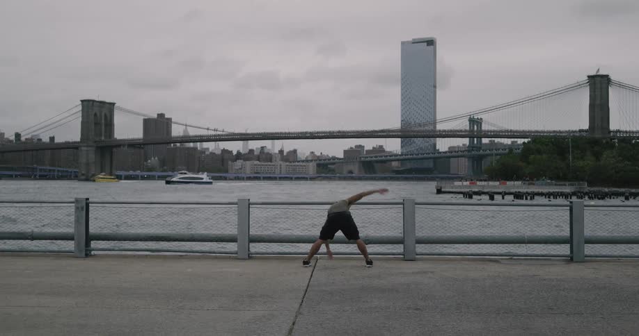 A man exercises on a sidewalk in front of a river across from a city skyline