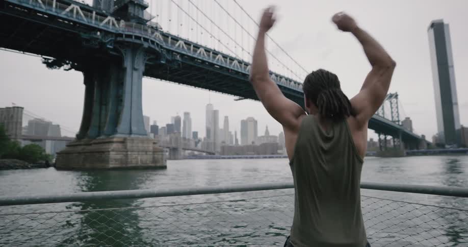 A man jogs to a corner on a pier and then does jumping jacks while looking out at the city harbor bridge