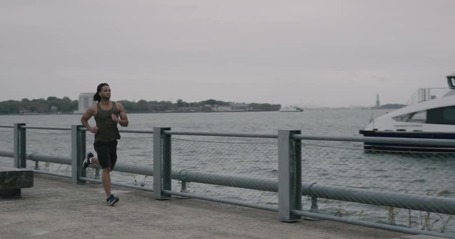 A muscular young male runs along a waterfront path that is across the water to a densely packed metropolitan city