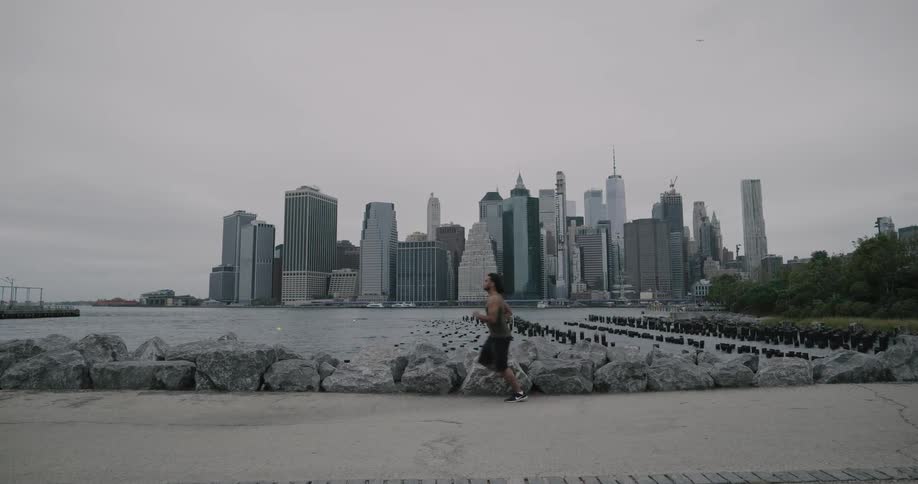 A young man jogs along a path at the edge of a body of water with a large metropolis on the other side