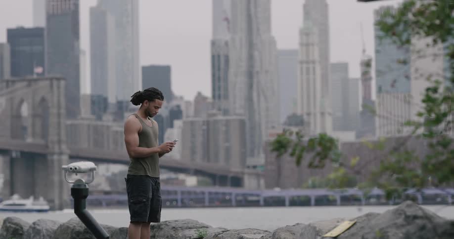 Black man stands on the promenade and uses smartphone next to the river across the new york city and brooklyn bridge on a cloudy day