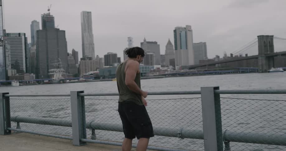 A man does jumping jacks and jogs in place on a pier while looking out at the harbor and city