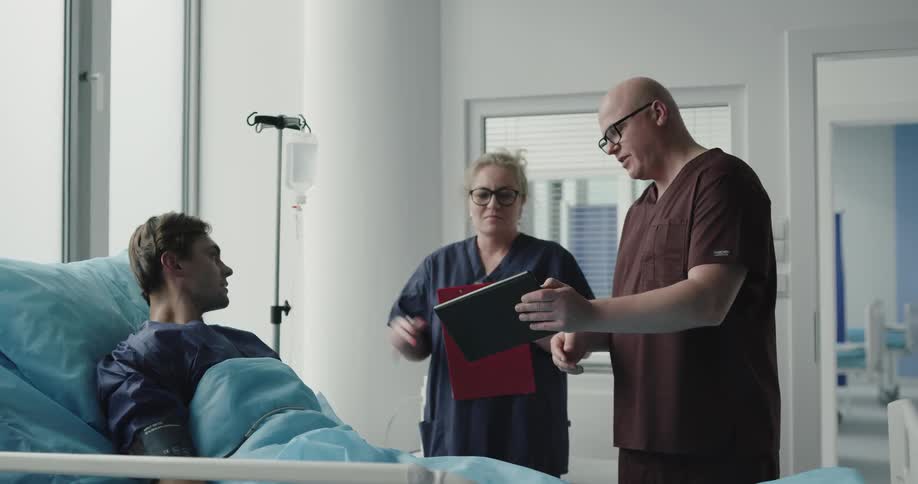 Two medical professionals walk into a room to check on a younger male patient who is sitting upright in a hospital bed