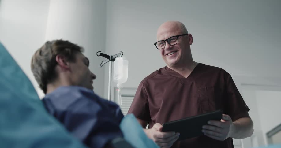 A nurse holds a tablet and talks to the patient in the bed