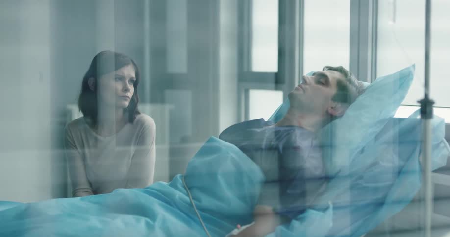A patient lays in a hospital bed hooked up to equipment while a loved one waits in a chair next to the bed