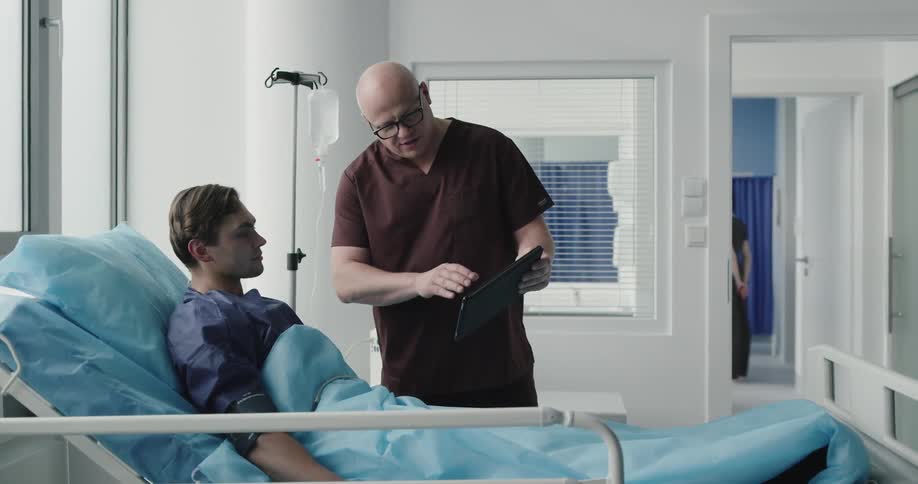 A doctor holds a tablet and talks about what it says to a patient laying in bed