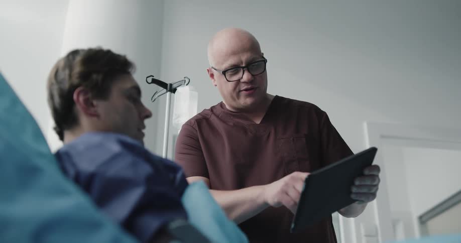 A man in a hospital bed being having something on a tablet being shown and explained to him by a male nurse