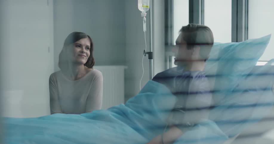 A man in a hospital bed is speaking with a woman seated by the bed