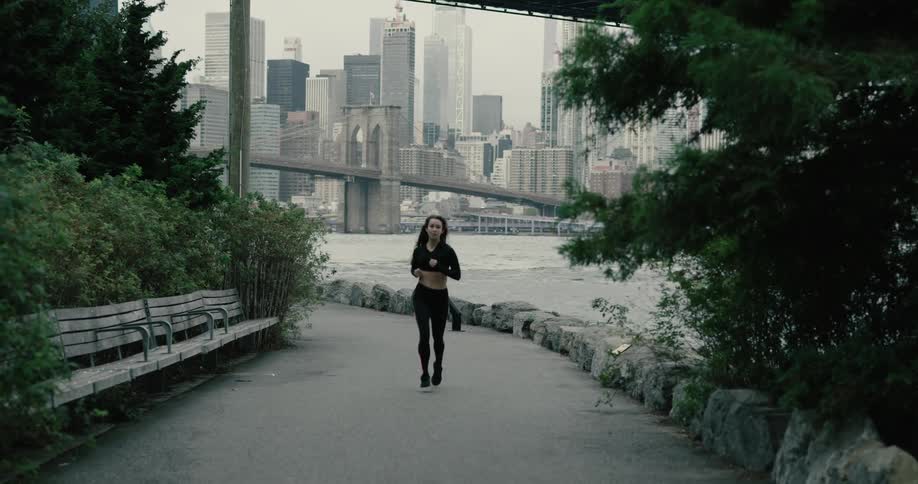 Woman runs along on the promenade next to the river across the new york city and brooklyn bridge on a cloudy day