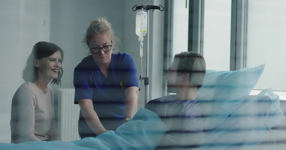 A man in a hospital bed talking with a woman while a nurse adjusts his iv drip