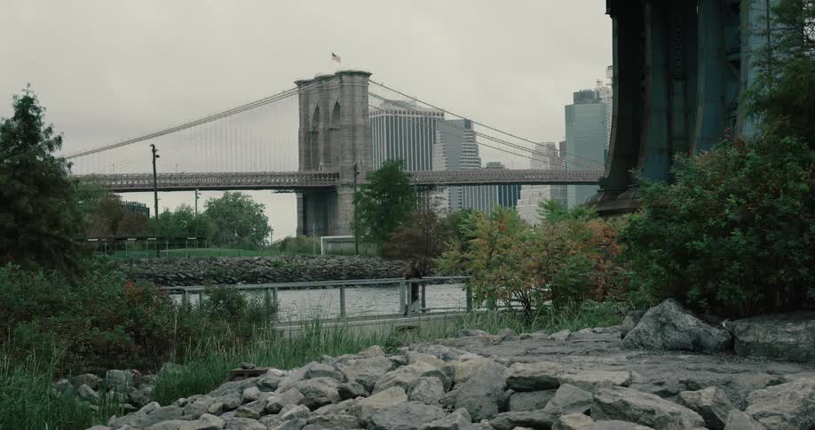 A woman jogs down a path in a park next to the brooklyn bridge on a cloudy day