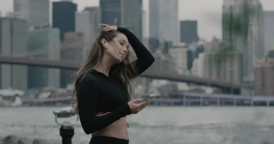 Woman stands on the promenade and uses smartphone next to the river across manhattan bridge and new york city on a cloudy day