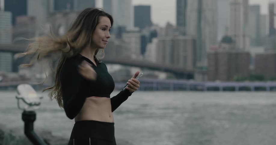 Woman runs along on the promenade and takes a break to use smartphone next to the sea across the city on a cloudy day