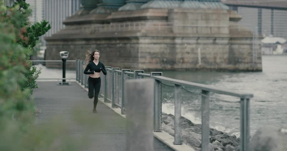 A woman jogs on a path while waves crash against the rocks at the base of a city harbor bridge