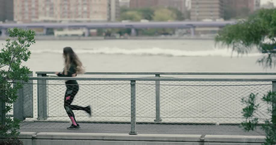 A woman in yoga pants and a crop top jogs next to a river as a boat floats by