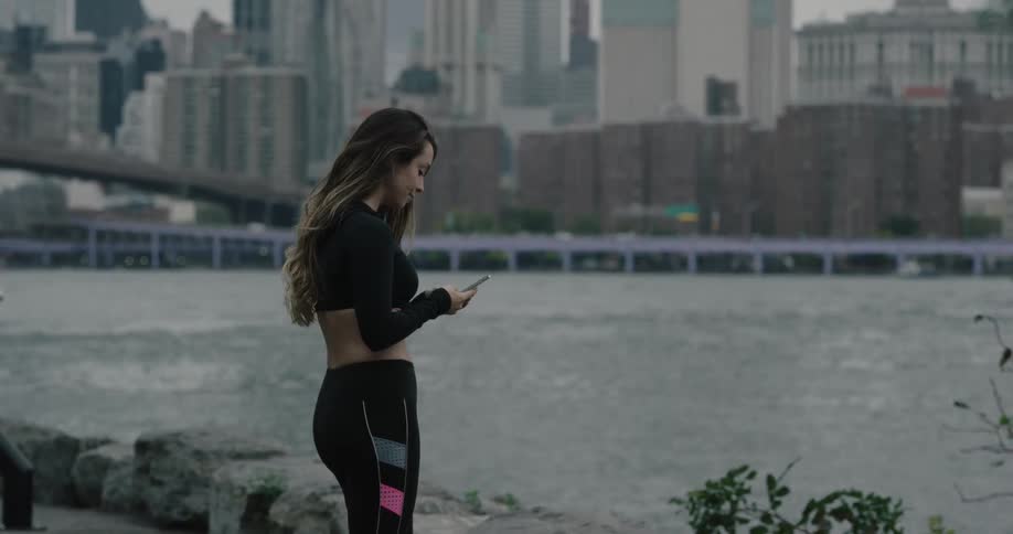 Woman holding smartphone runs along on the promenade and takes a break to wear bluetooth earphones next to the sea across the city on a cloudy day